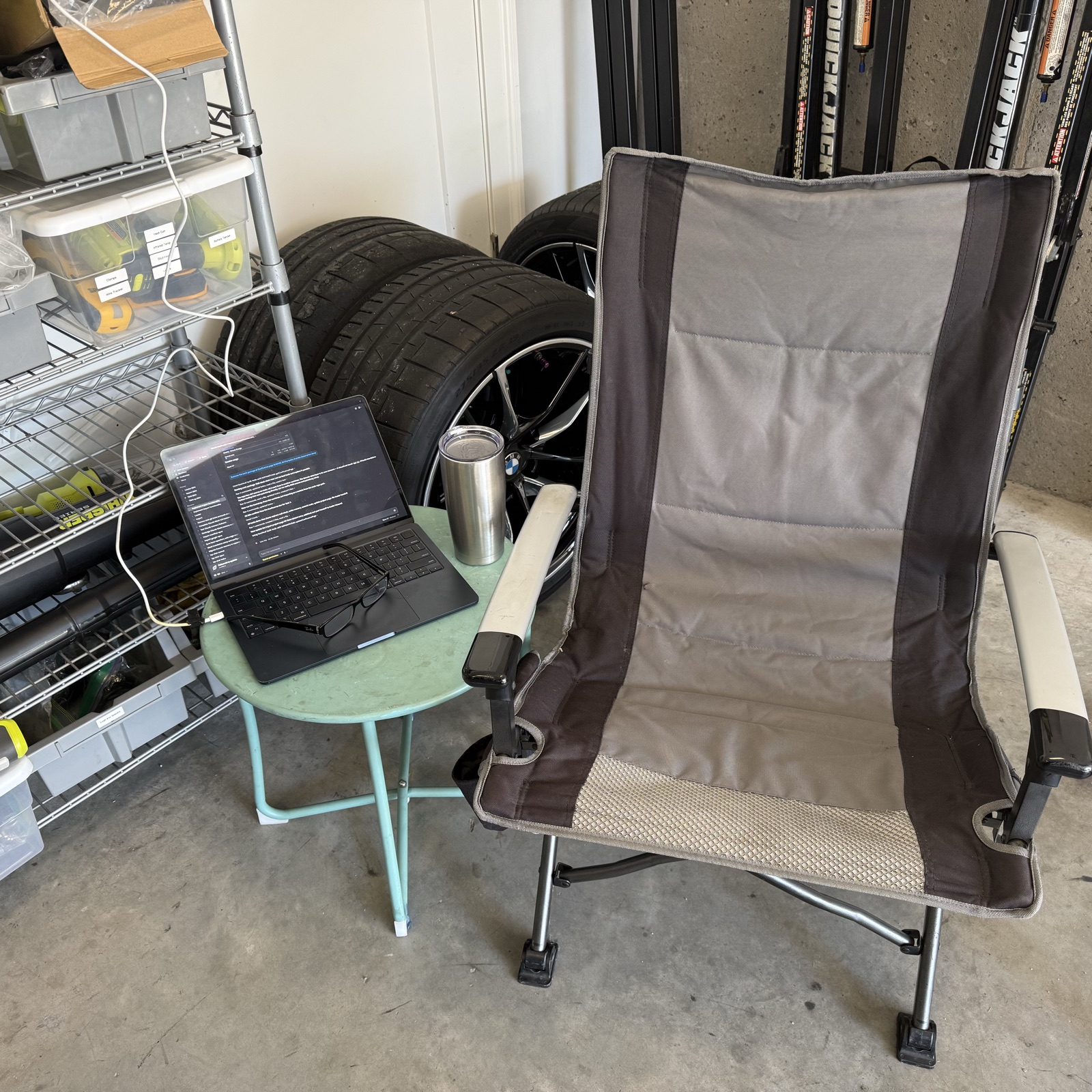 A laptop on a small green table in a garage, with a stack of BMW track wheels and a folding chair next to it.